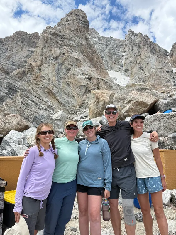 Five people stand arm in arm at a high camp beneath rocky peaks.