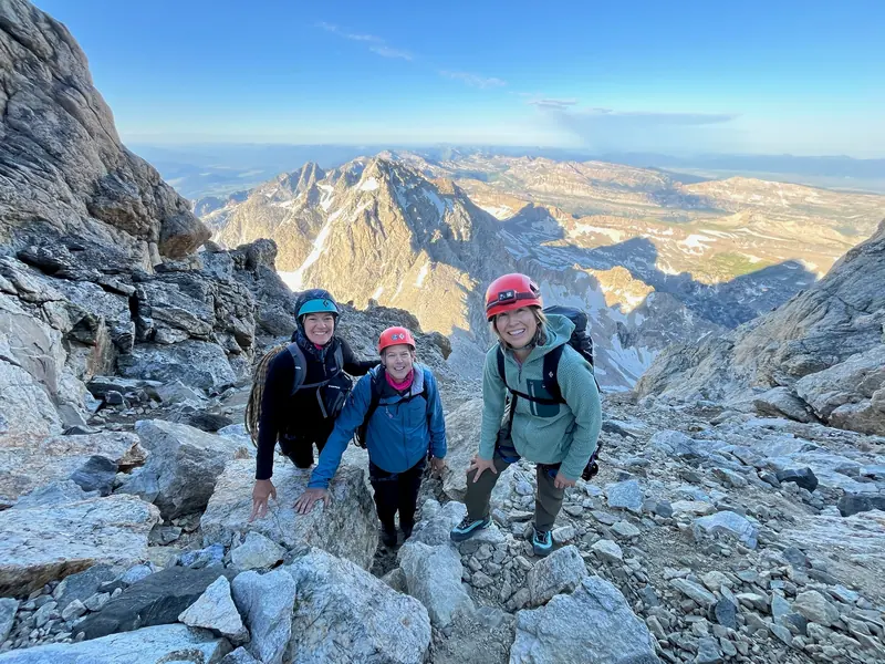 Three climbers in helmets smile on a rocky slope at sunrise, with snow-streaked peaks behind them.