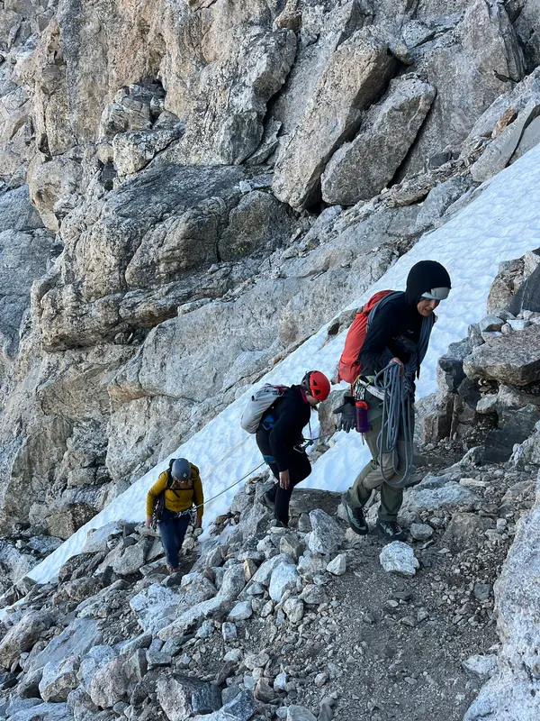 Three roped climbers cross rocky ground below a patch of snow.