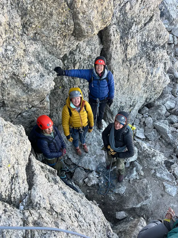 Four climbers in helmets and warm jackets look up from a notch in the rock.
