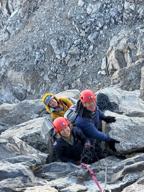Three climbers in helmets smile up at the camera from a gap in the rock.