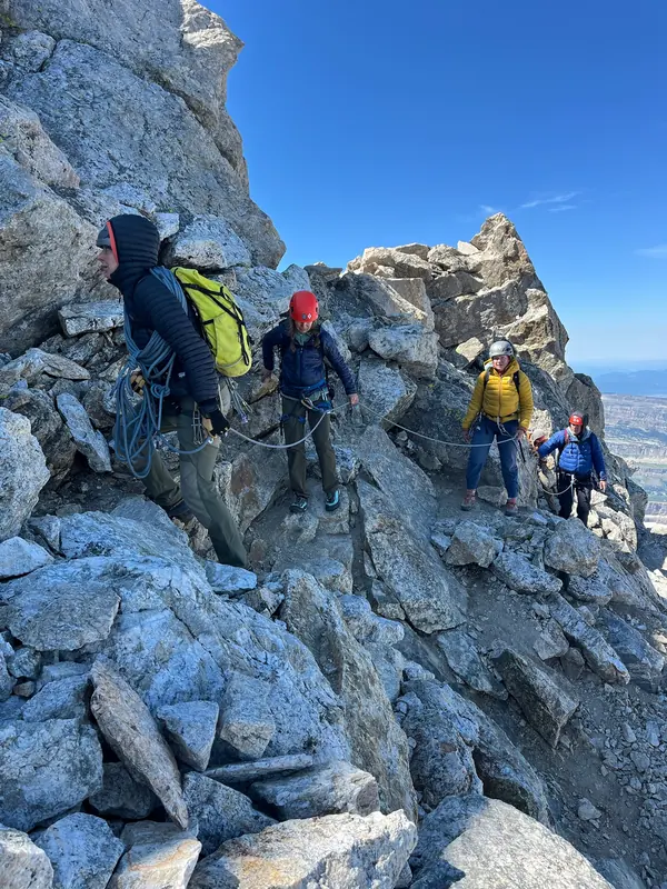 A roped team crosses a rocky ledge under a clear blue sky.