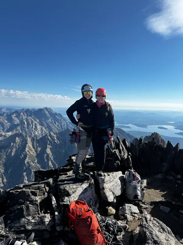 Two climbers stand side by side on the summit, with a lake and distant ranges far below.