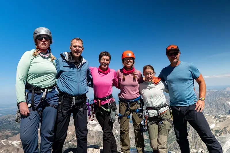 Six climbers stand arm in arm on the summit, with mountains stretching out behind them.