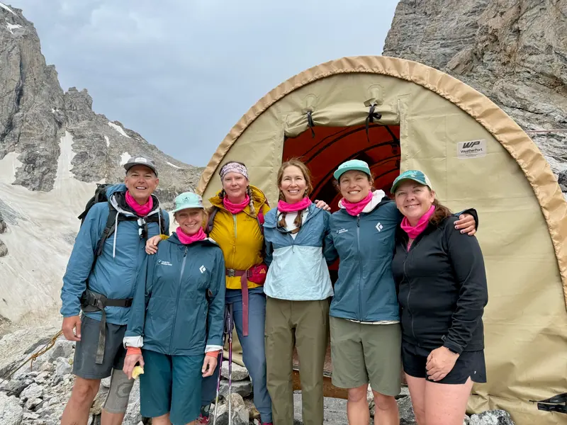 Six people in matching pink neck gaiters stand smiling in front of a domed hut high on the mountain.