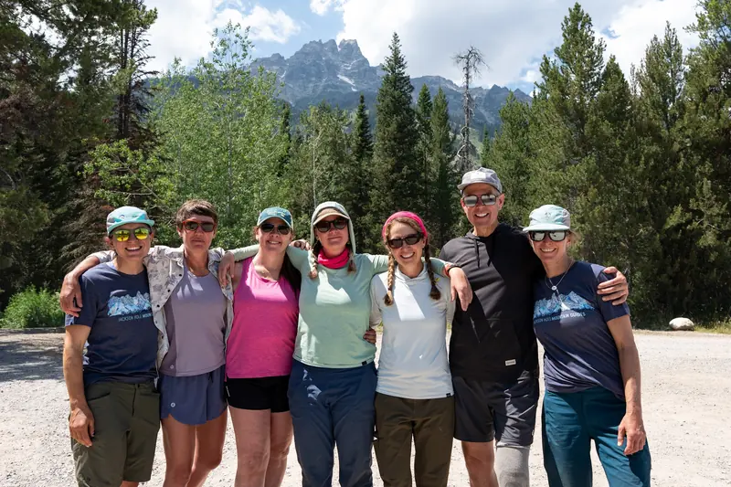 Seven people stand arm in arm at the trailhead, with the Tetons rising above the trees behind them.