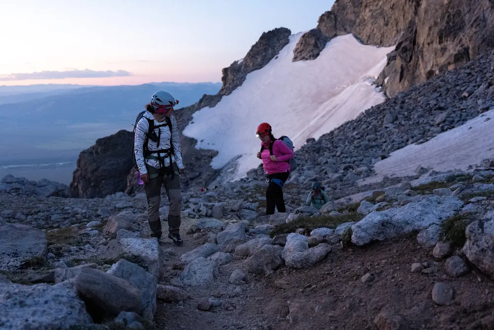 Two climbers on a rocky trail at dawn, a snowfield and cliffs behind them and the valley far below in pink light.